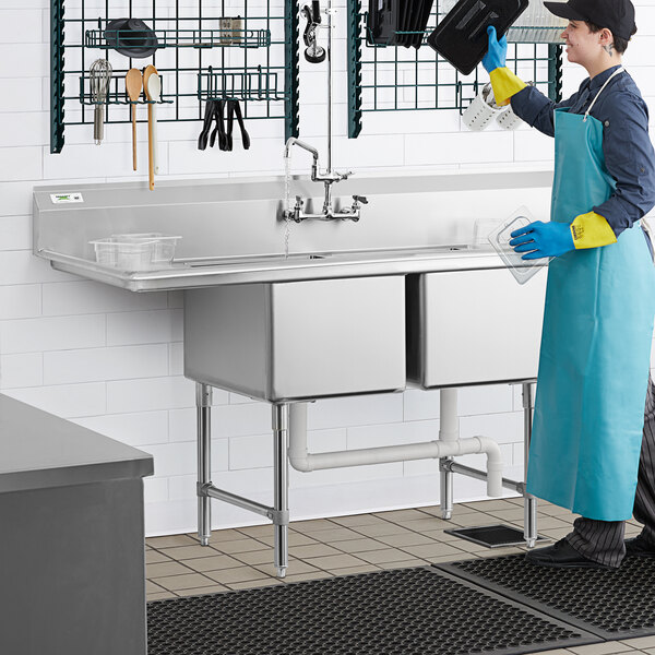 A man in a blue apron and yellow gloves using a Regency stainless steel two compartment sink in a professional kitchen.
