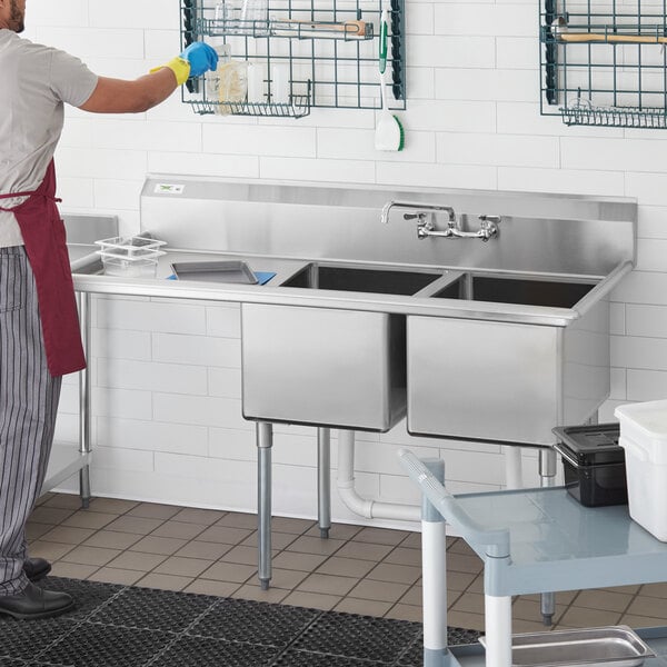 A man in a white apron washing dishes in a Regency two compartment sink with a left drainboard.