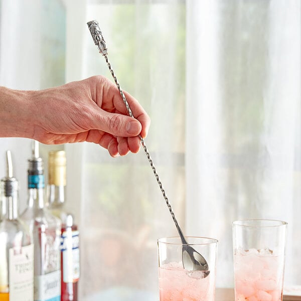 A hand using an Acopa silver bar spoon with tiki end to stir a pink cocktail.