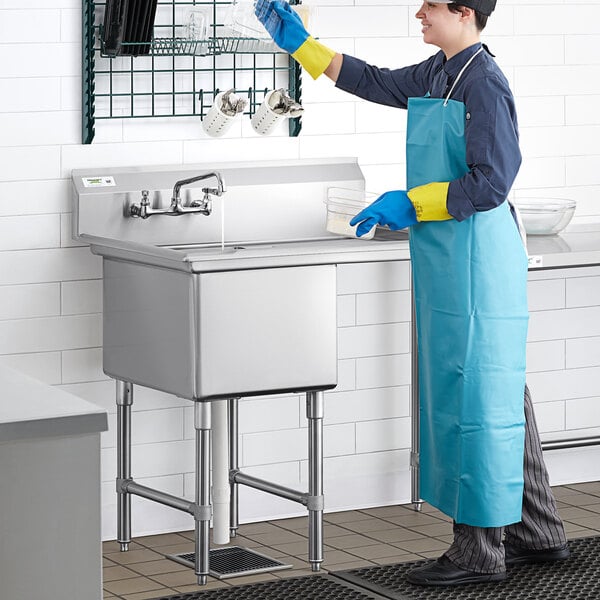 A man in a blue apron and gloves cleaning a Regency stainless steel commercial sink.