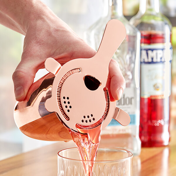 A hand using an Acopa copper Hawthorne strainer to pour a drink from a shaker into a glass.