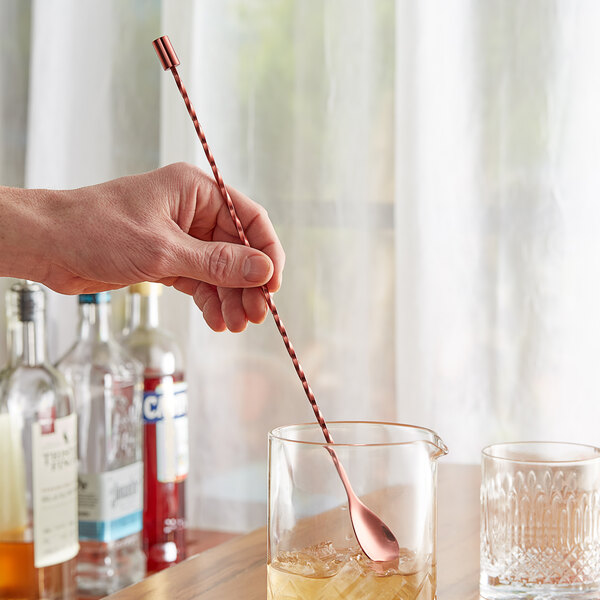 A hand using an Acopa copper bar spoon to stir a drink with ice.