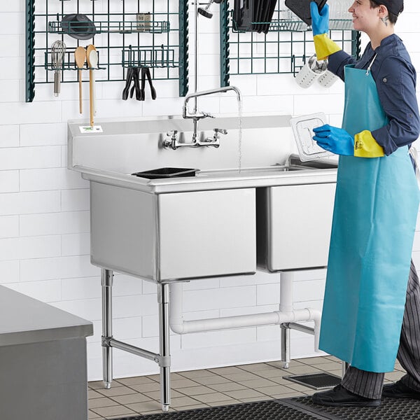 A woman in a blue apron washing dishes in a Regency two compartment commercial sink.