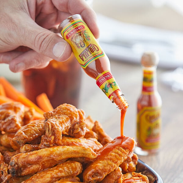 A hand pouring Cholula hot sauce onto a plate of wings.