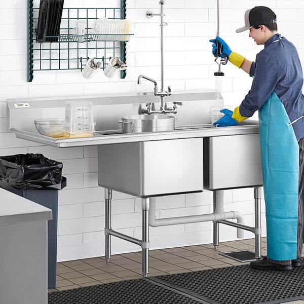 A person in a blue apron and gloves cleaning a Regency two compartment stainless steel sink.