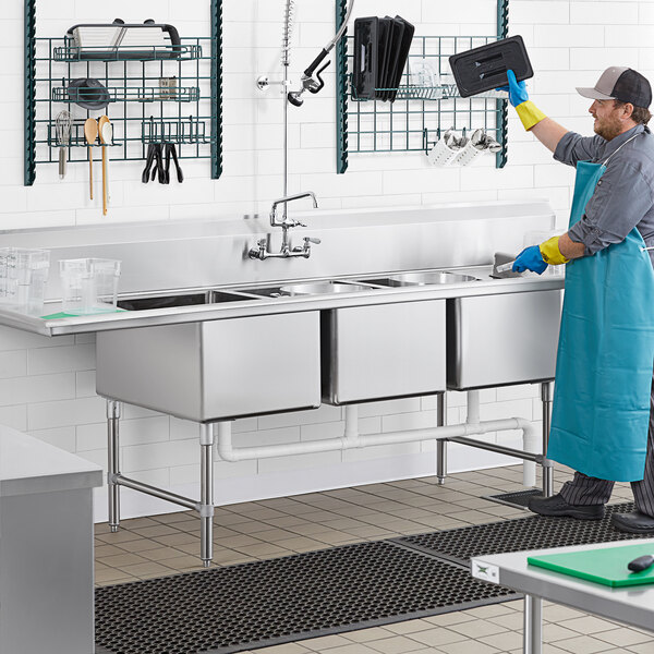 A man in a blue apron and gloves washing dishes in a Regency 3 compartment commercial sink.