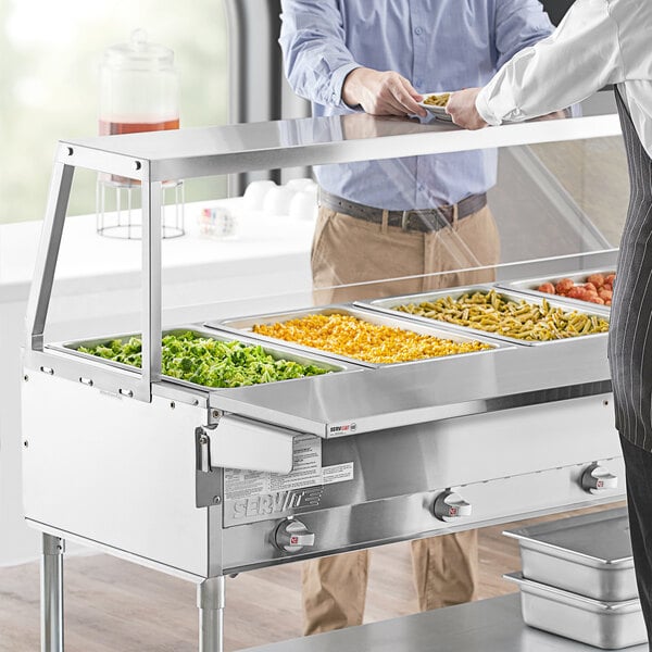 A man and woman standing in front of a buffet with a ServIt angled sneeze guard.