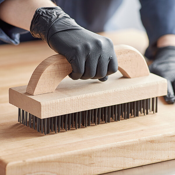 A person wearing black gloves uses a Choice wooden butcher block brush on a wooden cutting board.