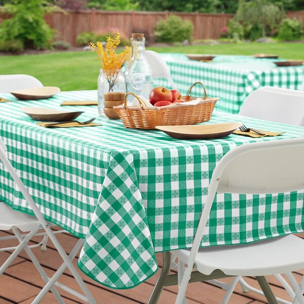 A picnic table with a green and white checkered vinyl tablecloth and a basket of fruit.