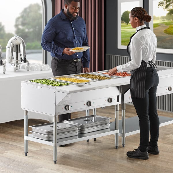 A man and woman serving food from a ServIt open well electric steam table at a hotel buffet.