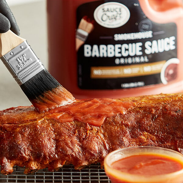 A hand using a paint brush to apply Sauce Craft Original BBQ Sauce to a piece of meat.