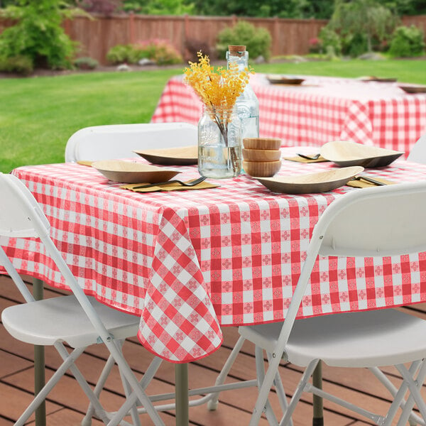 A table set with a red and white checkered vinyl tablecloth and food on it, with a yellow flower in a glass jar.