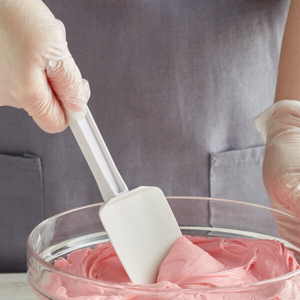 A person in gloves using a white Choice spoonula to mix pink frosting in a bowl.