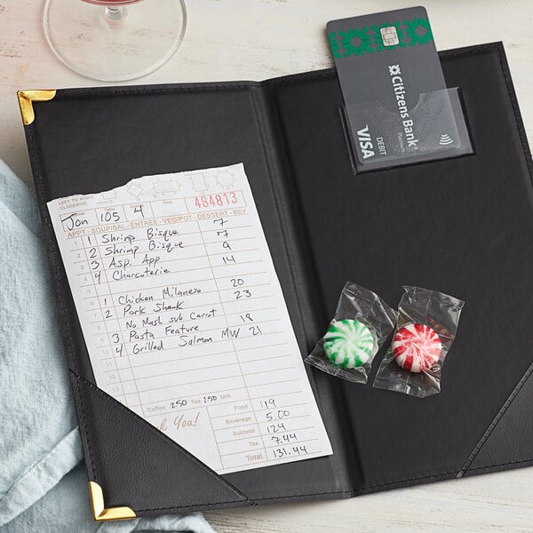A black leather Choice Guest Check Presenter with gold corners holding a receipt and a credit card on a table with candy and a drink.