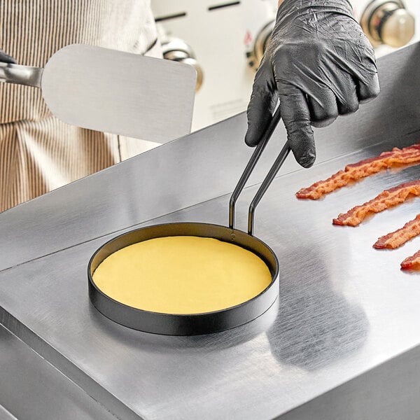 A person in black gloves using a Vigor egg ring to make bacon on a pan.