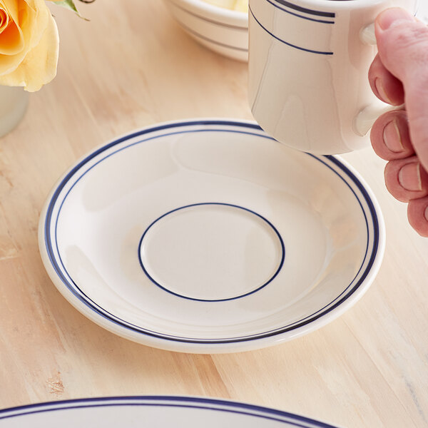 A hand holding an Acopa ivory stoneware cup over a saucer with blue trim.
