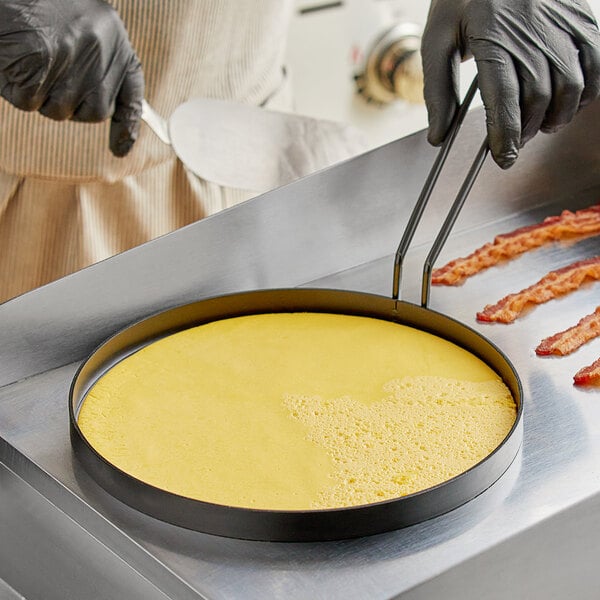 A person using a Vigor Non-Stick Egg Ring to cook bacon and eggs on a pan.