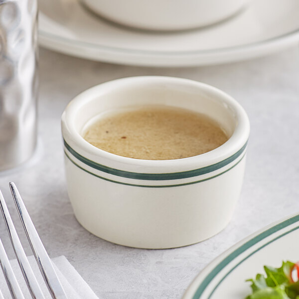 An Acopa ivory stoneware ramekin with green bands on a table with a bowl of brown liquid and a spoon.