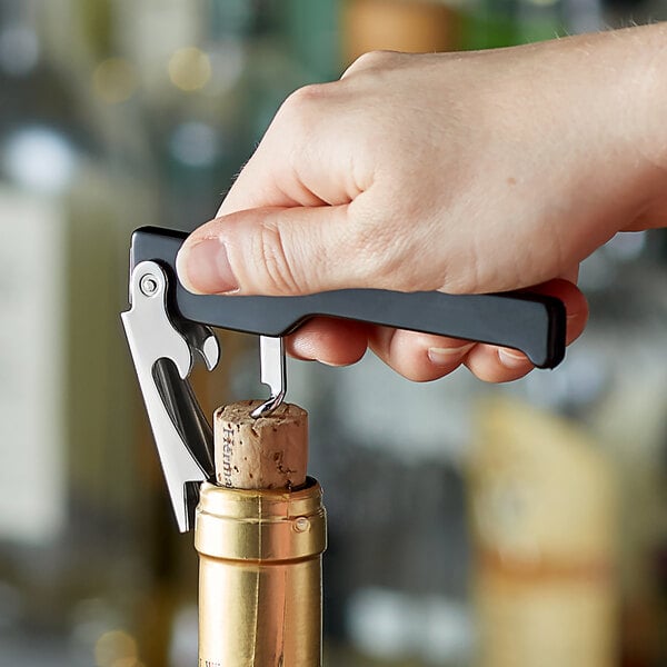 A hand using the Choice All-in-One Waiter Corkscrew to open a bottle of wine with a cork on a counter.