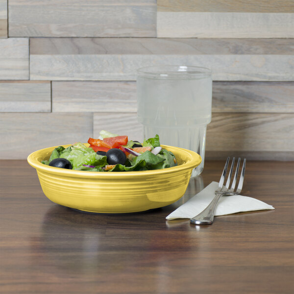 A Fiesta china bowl filled with salad on a table with a fork and a glass of water.