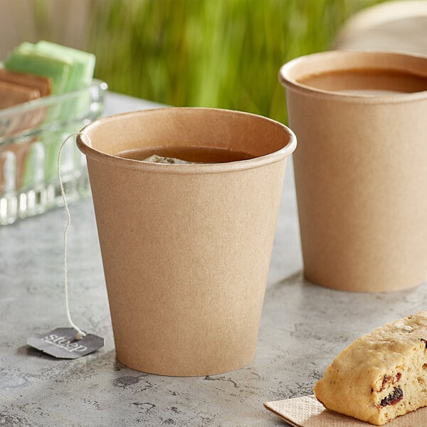 Two cups of coffee on a table with a pastry in between them in a Choice tall Kraft paper hot cup.