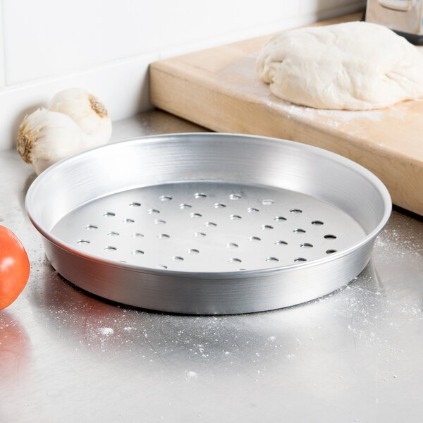 An American Metalcraft silver aluminum pizza pan with holes in it, next to dough and a tomato.