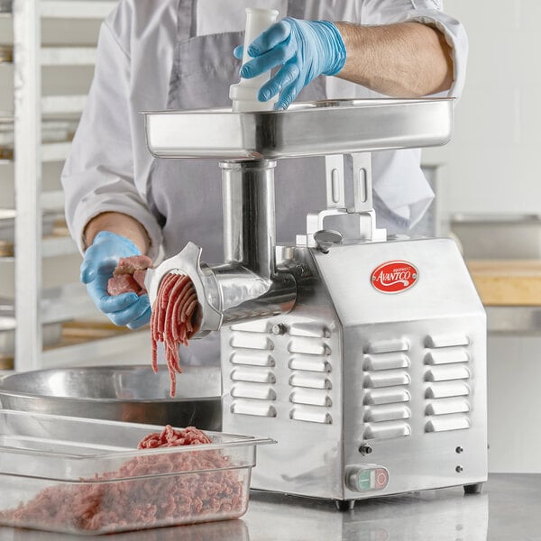 A person in a white coat and blue gloves using an Avantco meat grinder to grind meat.