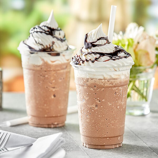 Two brown drinks in plastic cups with whipped cream and chocolate syrup on top.