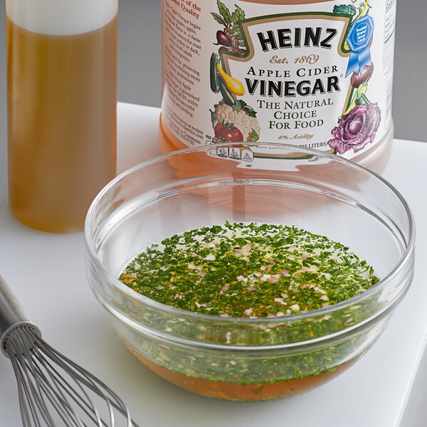 A bowl of green liquid next to a whisk and a bottle of Heinz Apple Cider Vinegar.