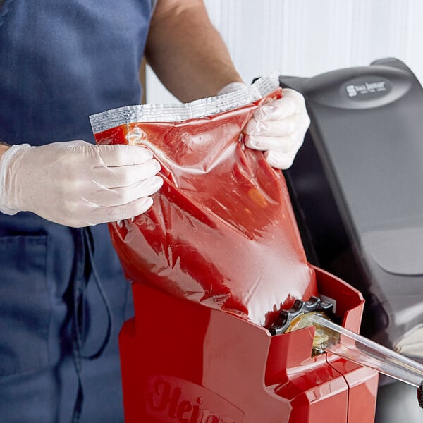 A person in gloves pouring red Heinz ketchup from a plastic bag into a machine.