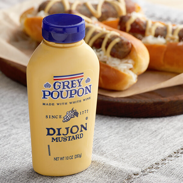 A close-up of a Grey Poupon Dijon Mustard squeeze bottle next to hot dogs.