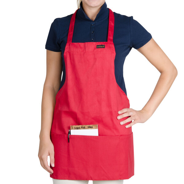 A woman wearing a red Chef Revival bib apron with a pocket in a professional kitchen.