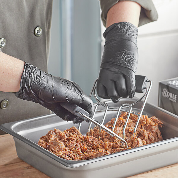 A person in Noble black gloves using a metal spatula to stir food in a container.