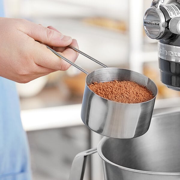 A person using an American Metalcraft stainless steel measuring cup to pour brown powder into a bowl.