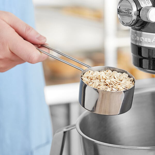 A person holding an American Metalcraft stainless steel measuring cup filled with oats.