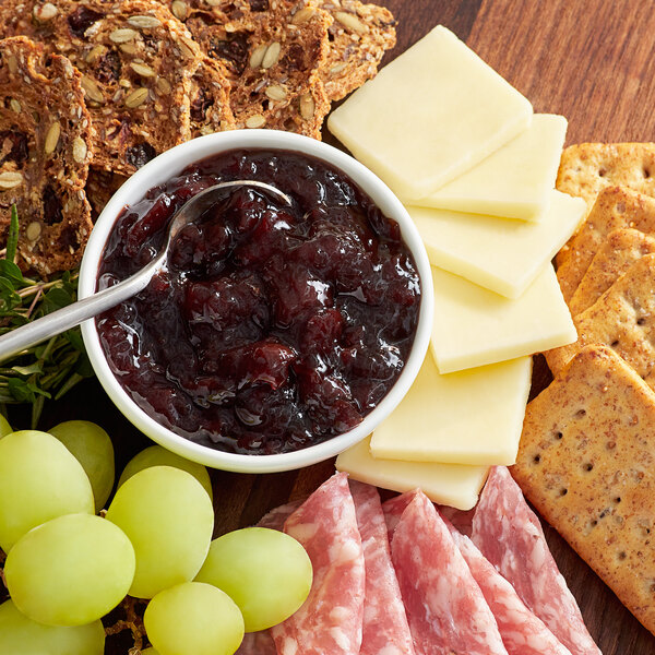 A wooden table with a bowl of Dalmatia Sour Cherry Spread, cheese, grapes, and crackers.