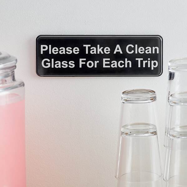 A black sign with white text that says "Please Take a Clean Glass for Each Trip" on a pink surface with a white stripe.