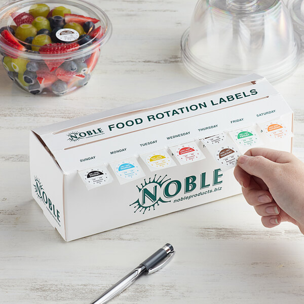 A hand holding a plastic container of Noble Products food rotation labels with day of the week clocks.