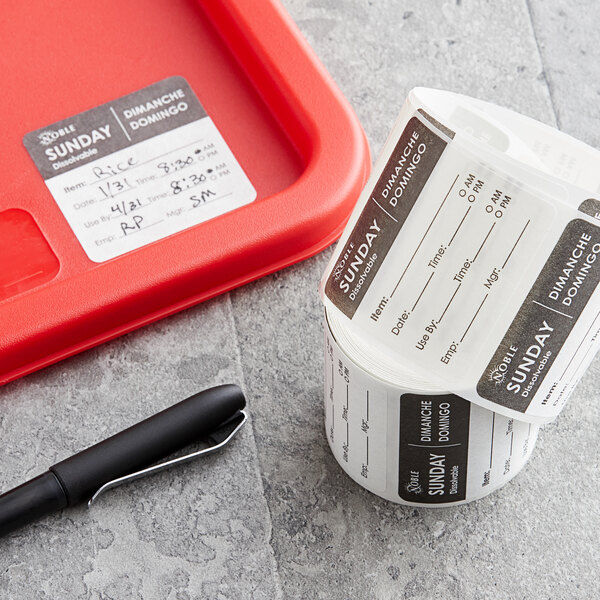 A roll of white and black Noble Products Sunday food labeling stickers on a grey surface next to a black pen.
