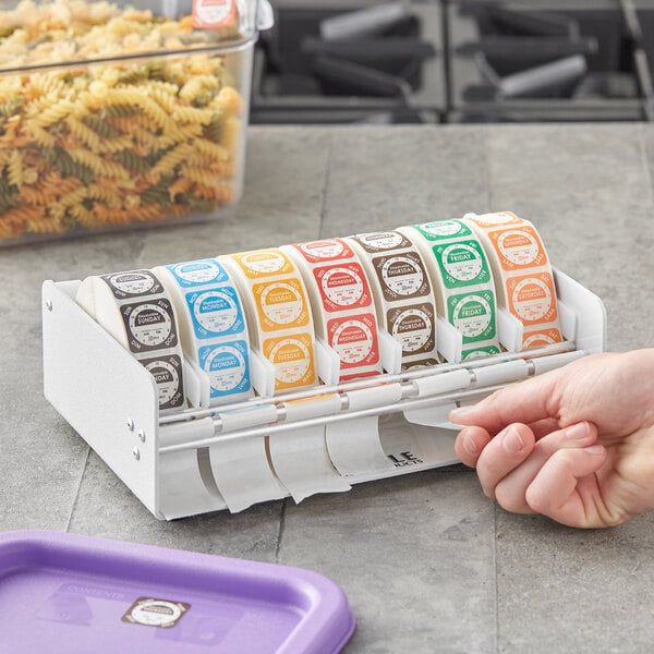 A hand using the Noble Products Elevated 7-Slot Dispenser to apply a white label with green and brown text to food on a purple tray.