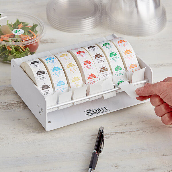 A hand using a dispenser to put a Noble Products day of the week label on a plastic container of salad.
