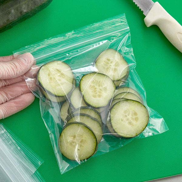 A gloved hand slices a cucumber and places a slice in a Clear Line plastic bag.