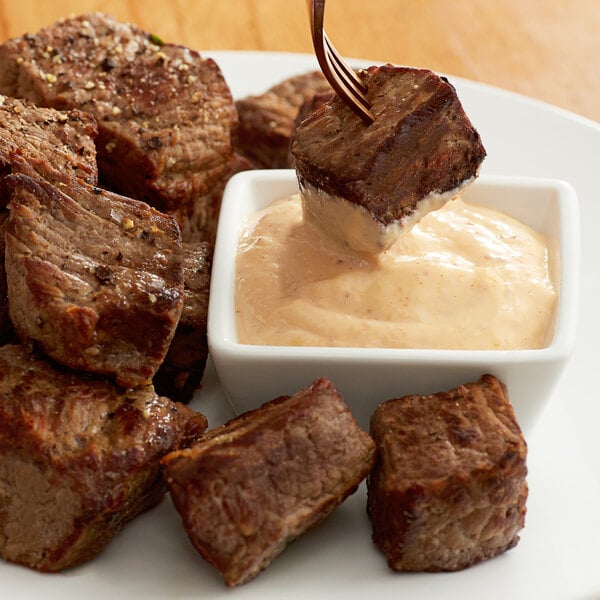A fork dipping steak into a white bowl of Grey Poupon Bistro Sauce.