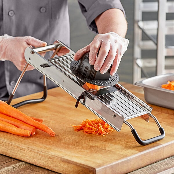 A person using a stainless steel mandoline to cut carrots on a cutting board.