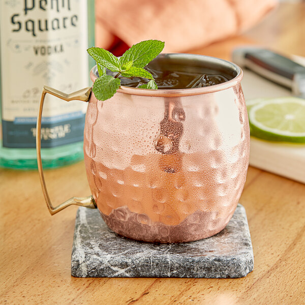 An Acopa Alchemy copper Moscow Mule mug with ice and mint leaves on a marble coaster.