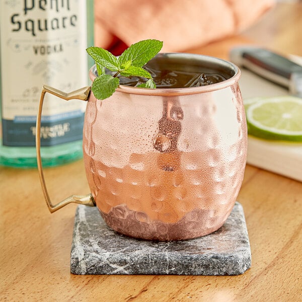 A Acopa hammered copper Moscow Mule mug on a marble coaster with ice and mint leaves.