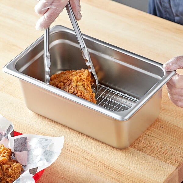 A person using tongs to cut up a chicken in a stainless steel steam table pan on a wood surface.