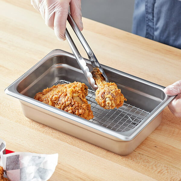 A person using tongs to remove fried chicken from a stainless steel tray.