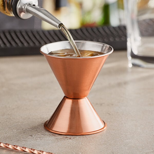 A close-up of a liquid being poured into a metal cup with a copper jigger on a table.