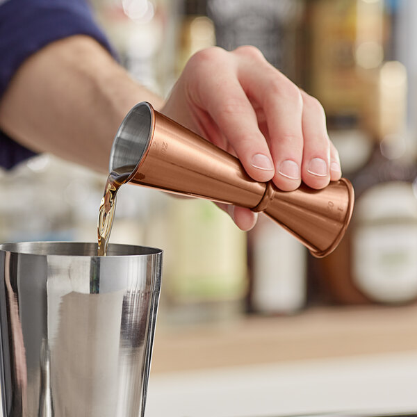 A person using an Acopa copper Japanese jigger to pour liquid into a metal cup.